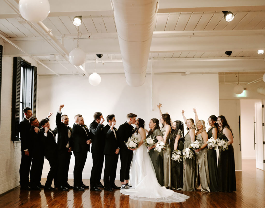 congdon yards silk room at the loft bridal party celebrating while bride and groom kiss